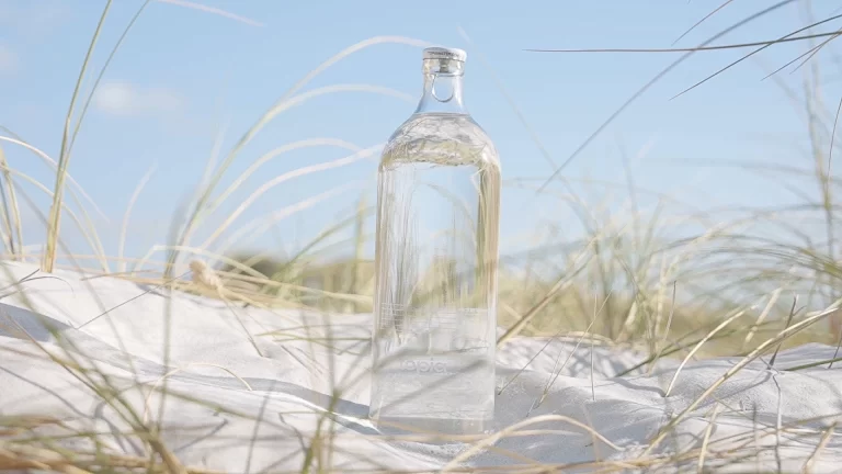 A clear bottle of sparkling water with a few bubbles sits on top of a sandy beach dune. The dune has green grass growing on it. [Topia, carbonated water, sparkling water]