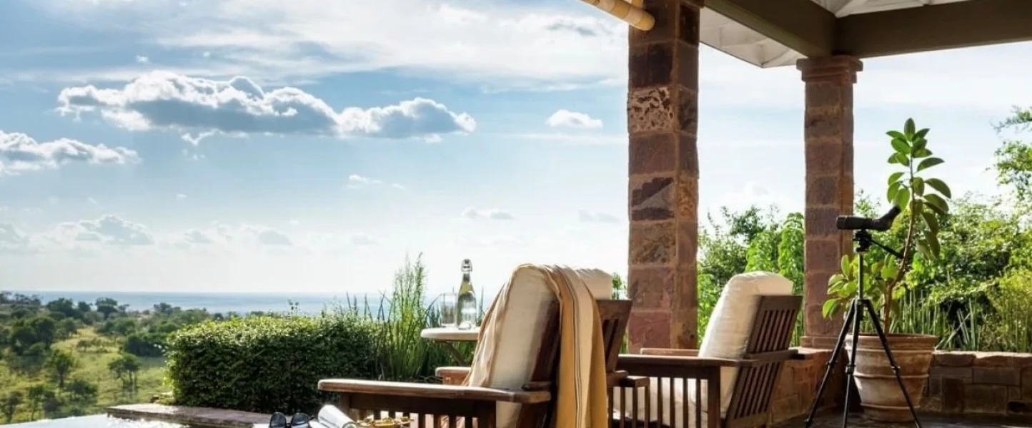 An expansive view from a luxurious safari lodge veranda overlooking a serene landscape, with a calm infinity pool blending into the distant horizon. Near the pool's edge, a pair of sunglasses and a transparent, reusable water bottle filled with fresh water rest on a woven wicker side table, suggesting preparedness for an adventure. Comfortable wooden armchairs with plush cushions invite relaxation, and a high-quality telescope mounted on a tripod promises up-close wildlife sightings, evoking a sense of anticipation for the day's safari explorations. The scene encapsulates a perfect blend of comfort, sustainability, and the thrill of the wild, highlighting the importance of staying hydrated with a reliable water bottle during such excursions.