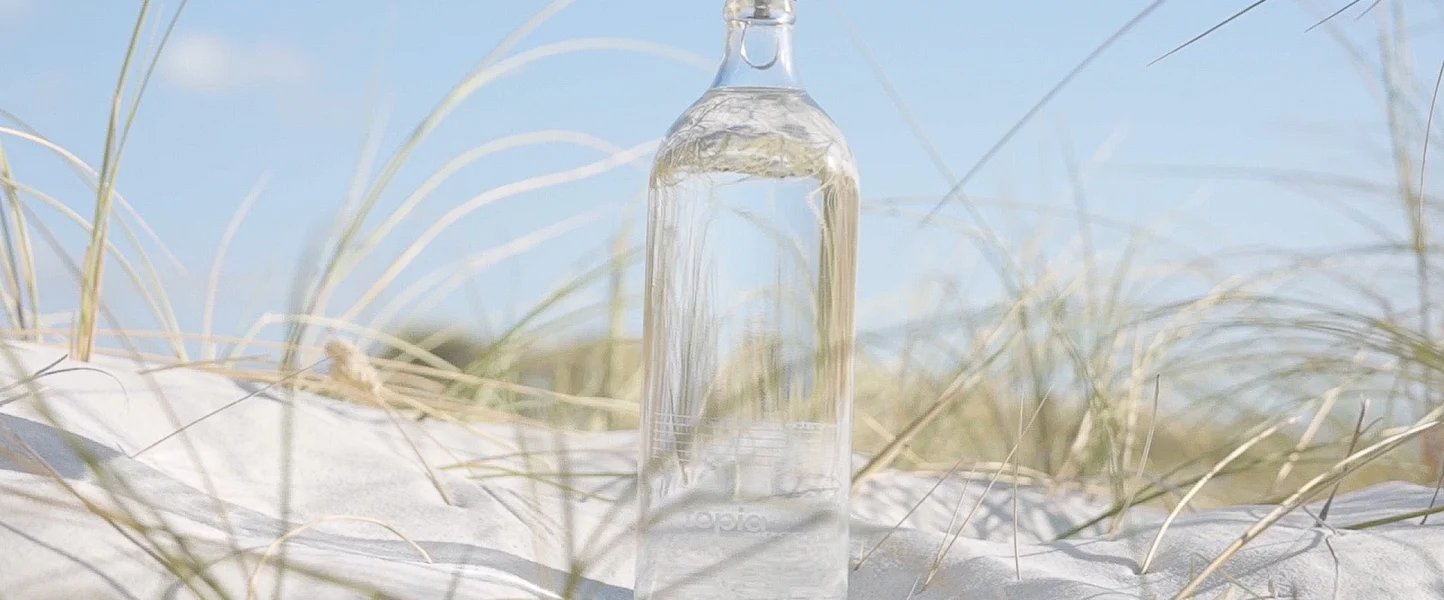 A clear bottle of sparkling water with a few bubbles sits on top of a sandy beach dune. The dune has green grass growing on it. [Topia, carbonated water, sparkling water]